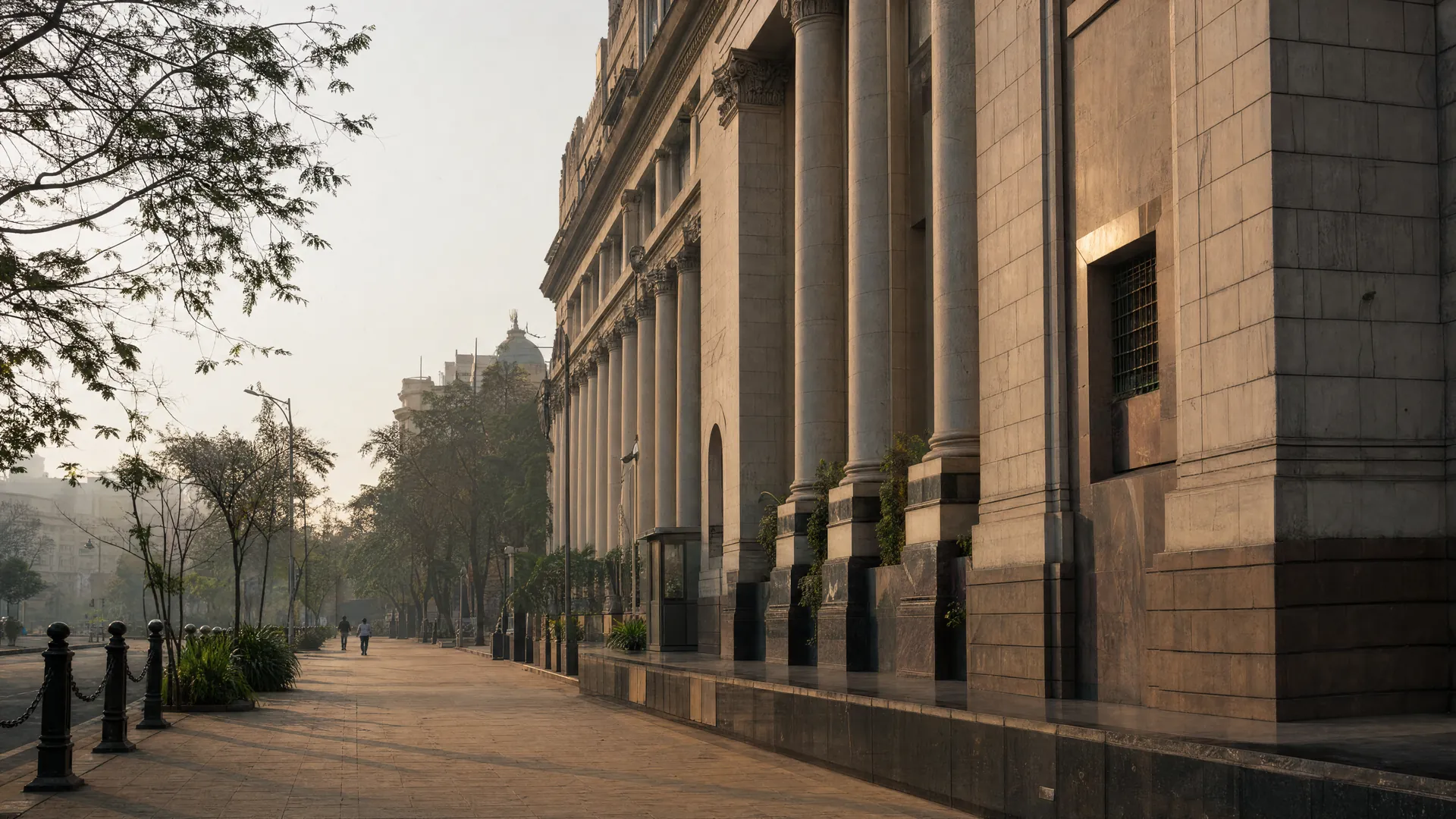 A stately stone central bank building in Mumbai at early morning, warm light on its columns, with a few distant pedestrians under a hazy sky.