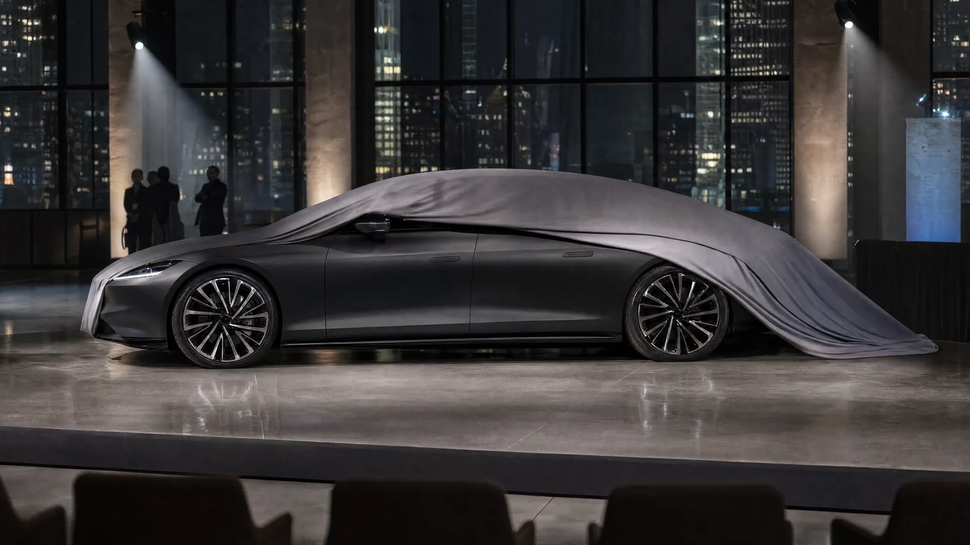 A sleek four-door electric car, partly under a gray cover, sits spotlit on a dark event stage with city lights glowing through tall windows.