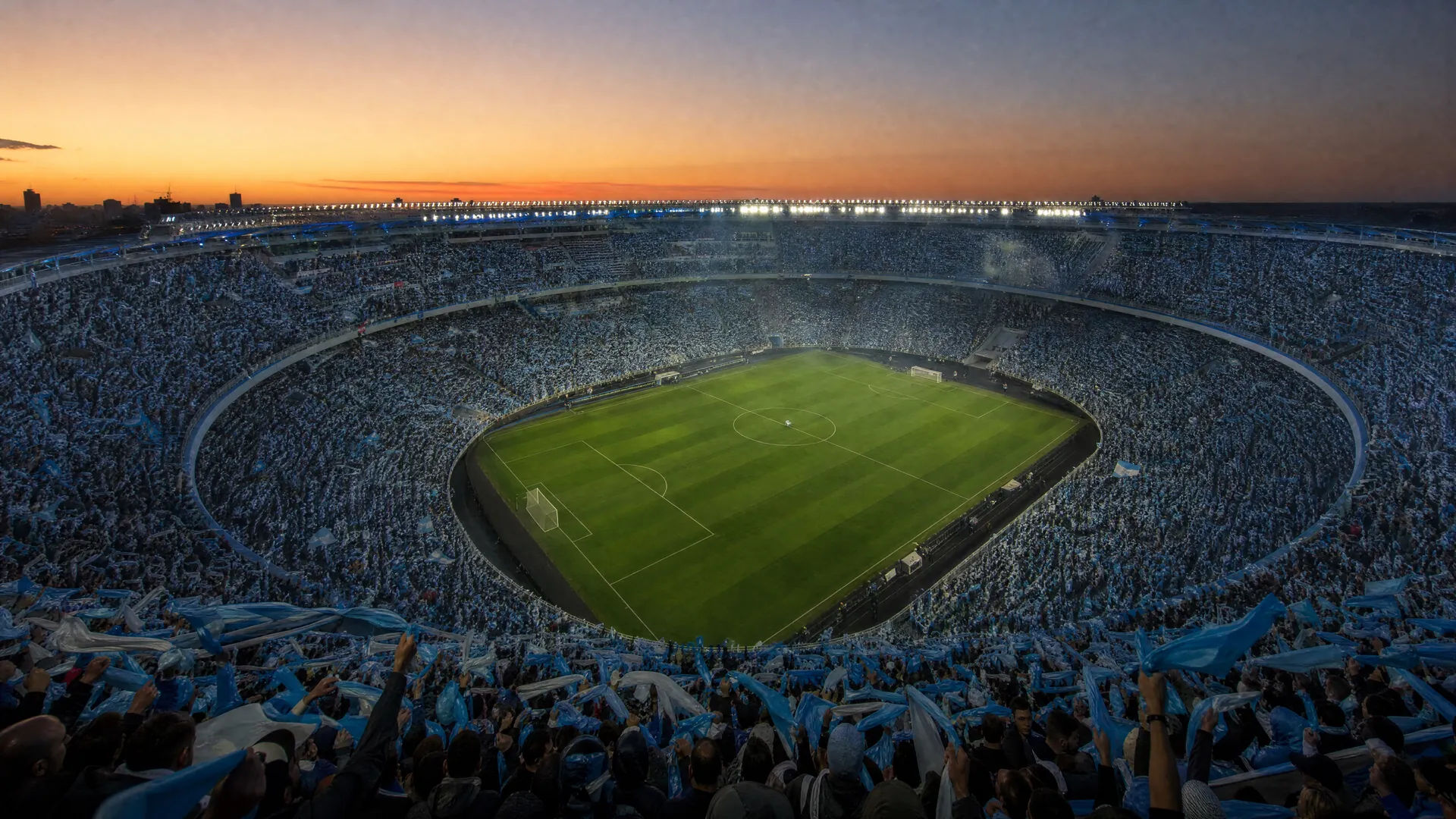 A floodlit stadium at dusk filled with distant fans in sky blue and white, a single soccer ball resting on the center spot of the pitch.