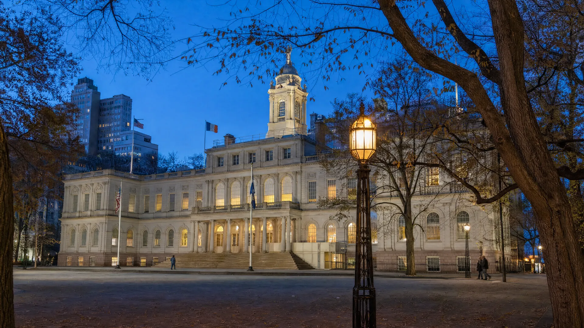 New York City Hall at dusk, its columned facade lit against a blue sky, with a few distant figures crossing the plaza under autumn trees.