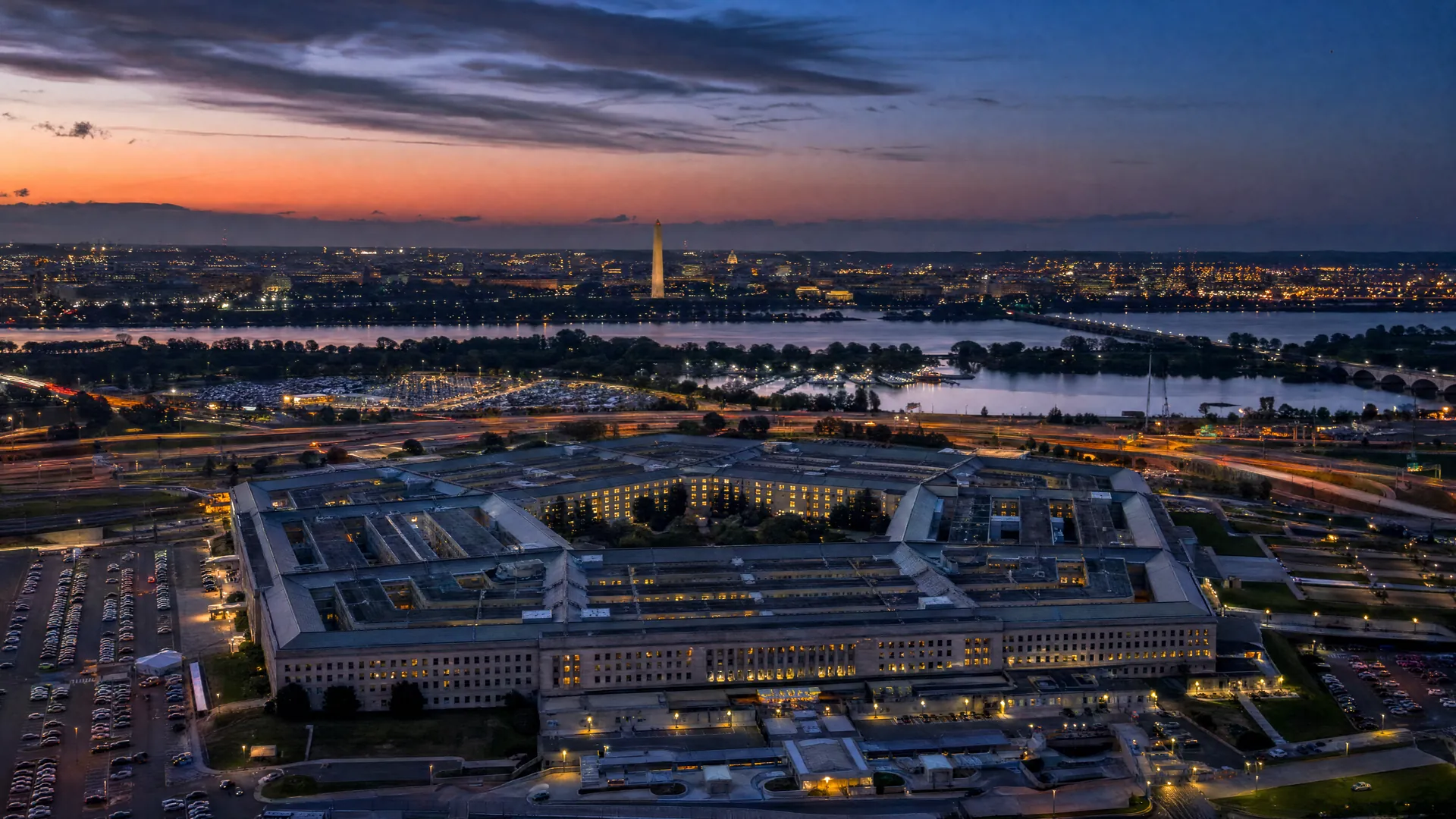 Aerial view of the Pentagon at dusk, a few windows lit, with the Potomac River and Washington skyline fading in the background.