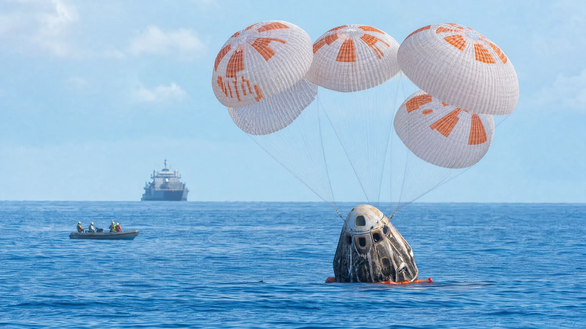 A white space capsule floats on calm ocean water beside settling orange-and-white parachutes, with a recovery ship on the horizon.