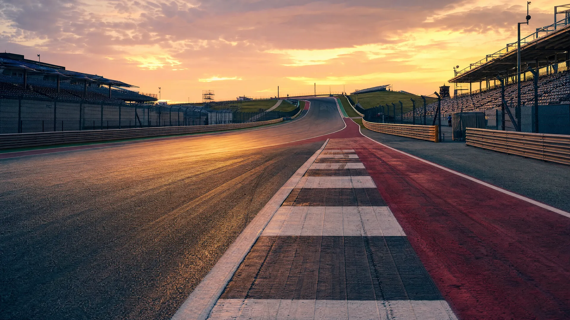 An empty racetrack straight and uphill first corner at golden dusk, with faint tire marks on the asphalt and empty grandstands under a pastel sky.
