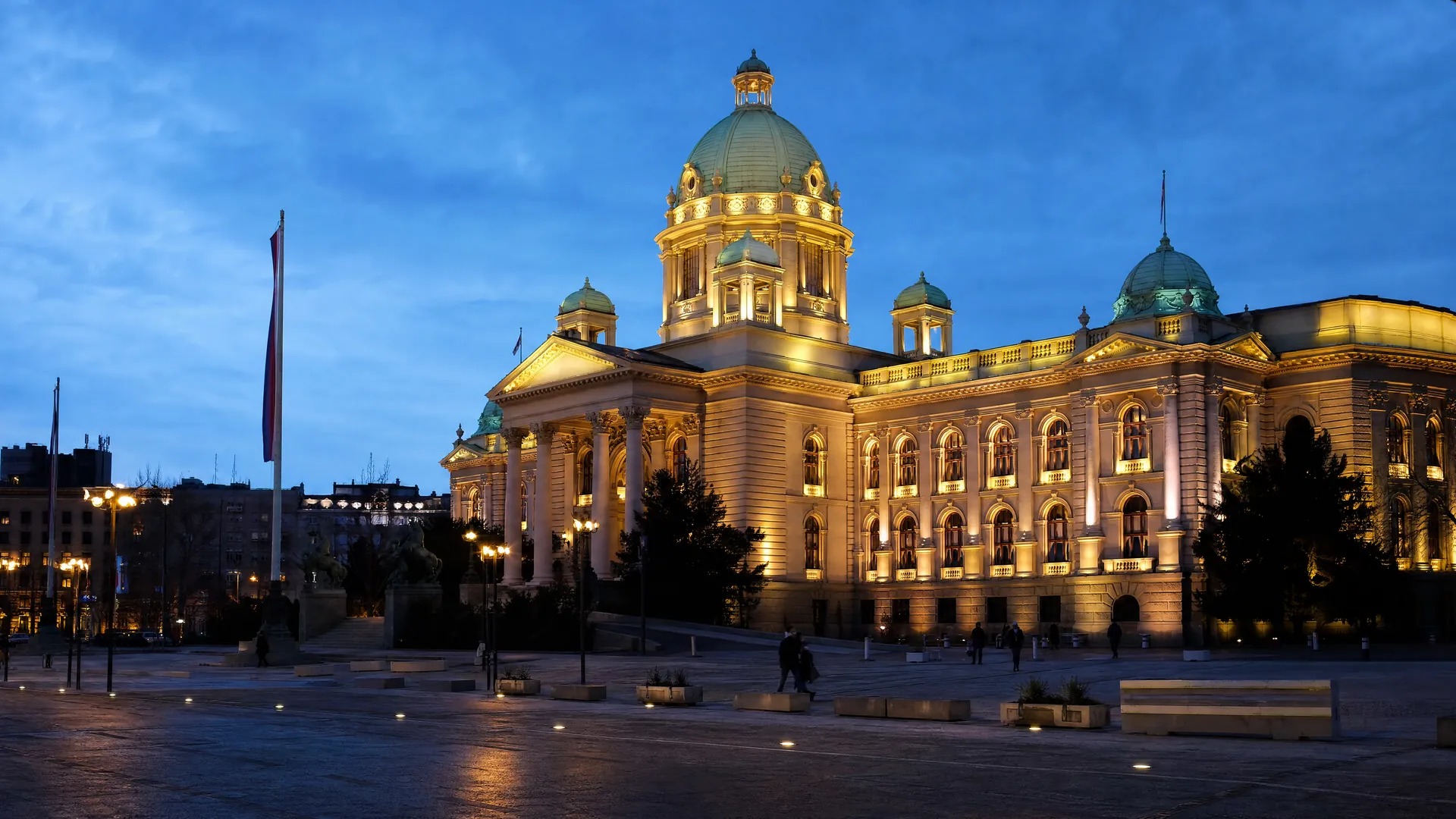 Serbia's National Assembly building in Belgrade at dusk, its domed facade lit warmly, with a few distant figures crossing the square.