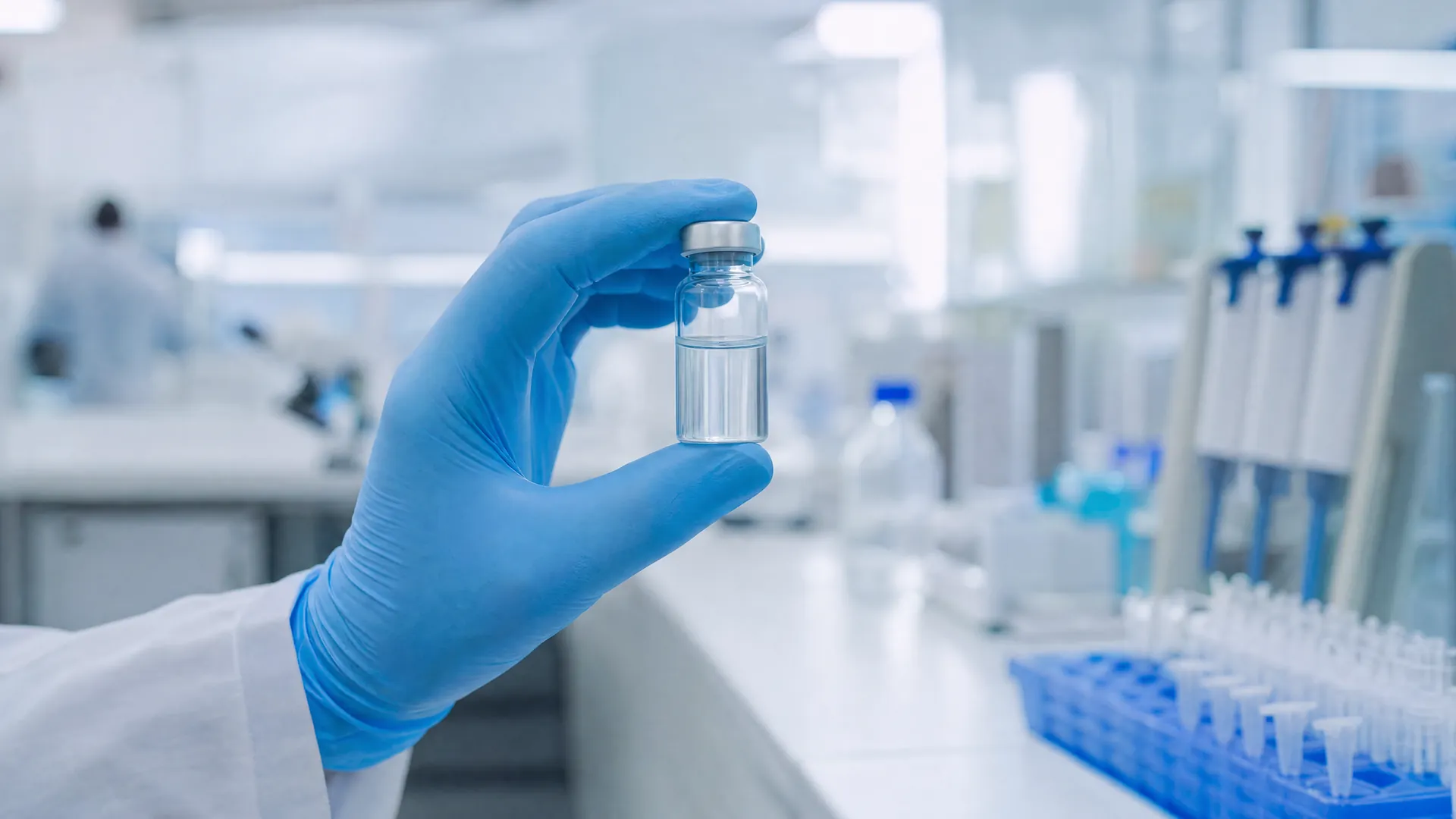 A gloved hand holds a small clear vaccine vial in a bright, modern laboratory with blurred equipment behind it.