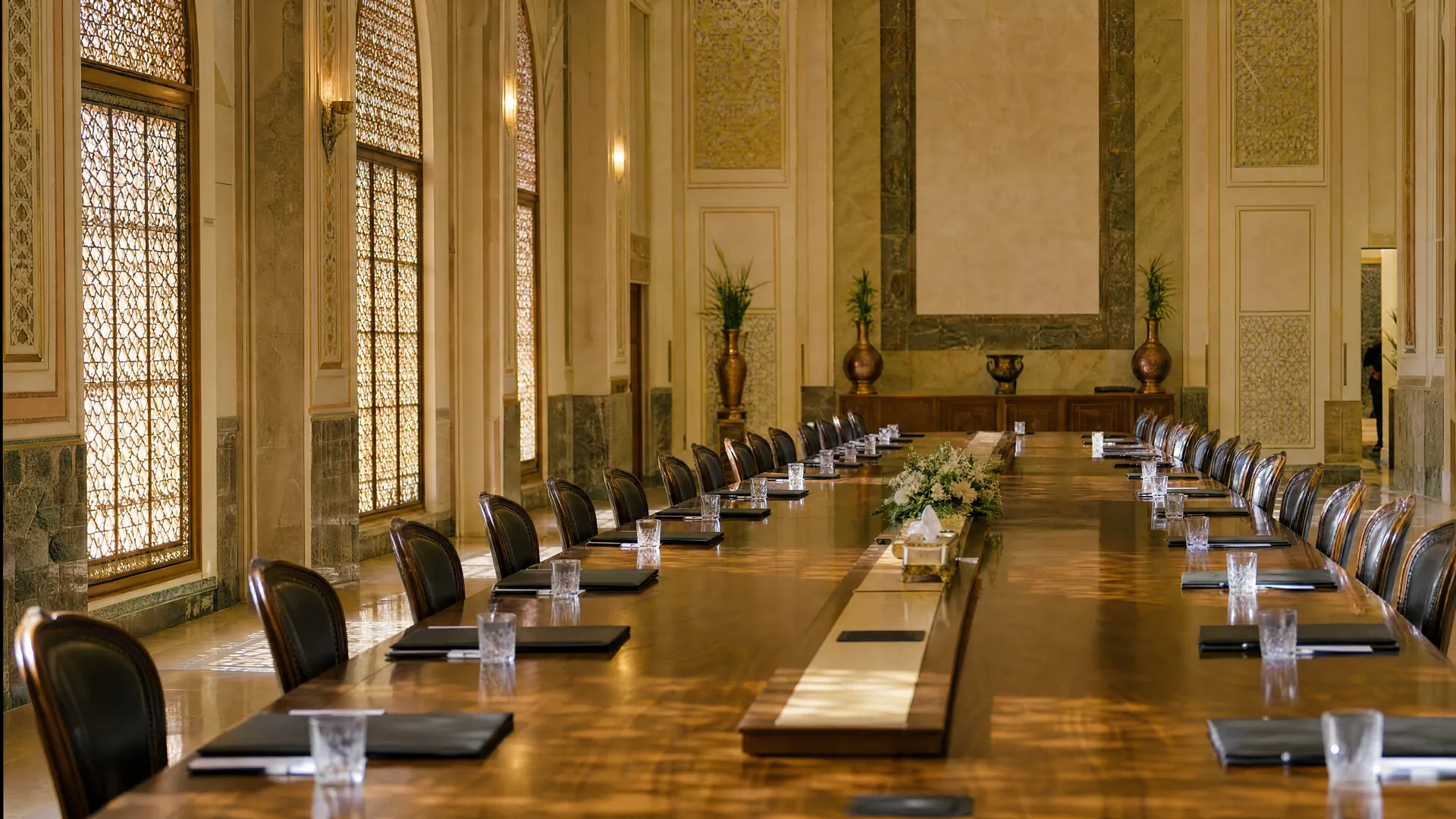 An empty, ornate conference hall with a long polished table, neat chairs and closed folders, lit by soft light through latticed windows.