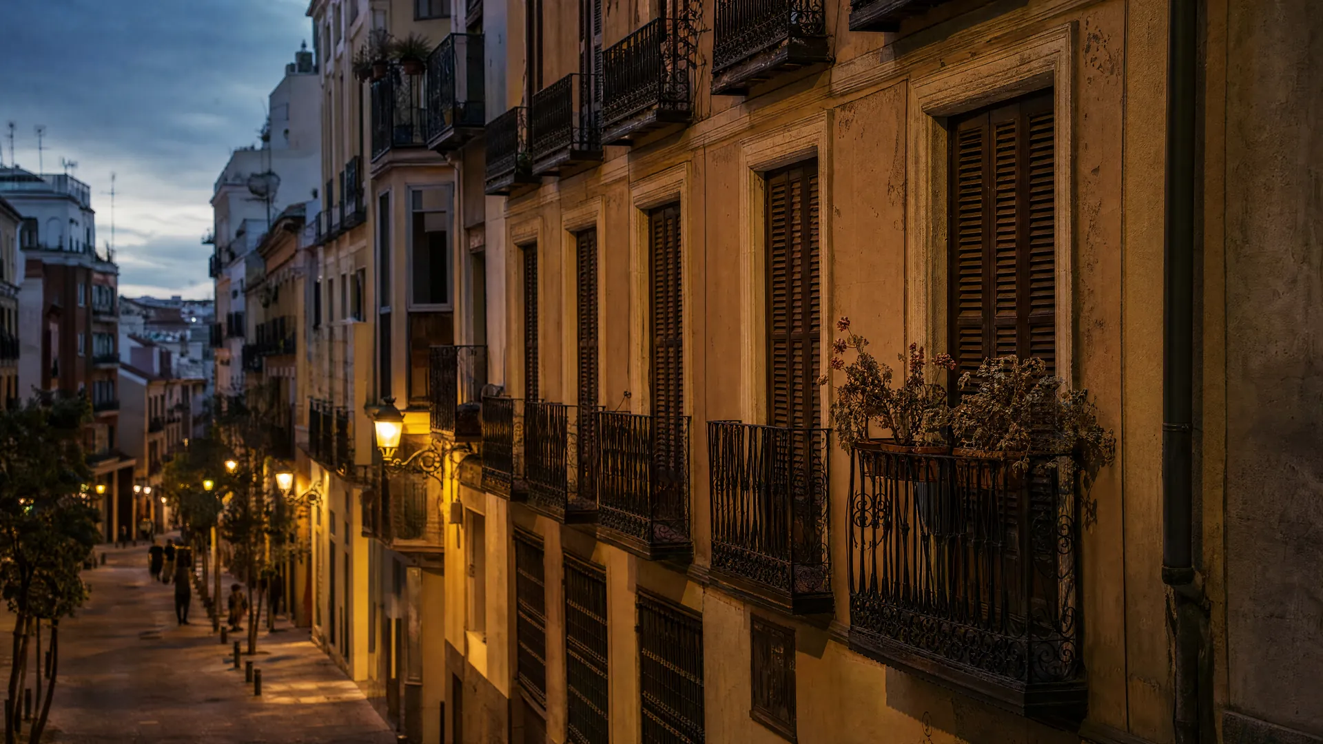 An old Madrid apartment building at dusk, shutters closed and wilting geraniums on a balcony, with distant pedestrians below.